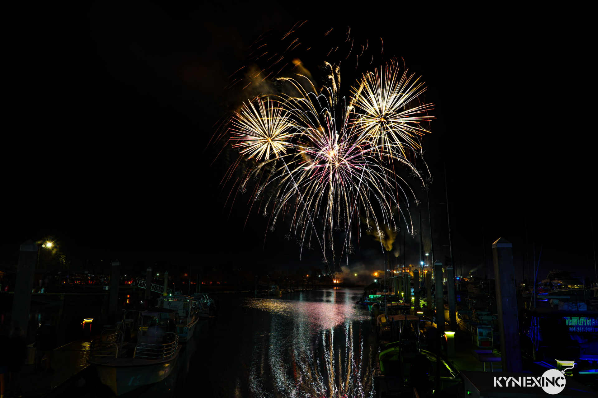 Fireworks bursting over the Amelia River with the Fernandina Beach skyline reflected in the water.